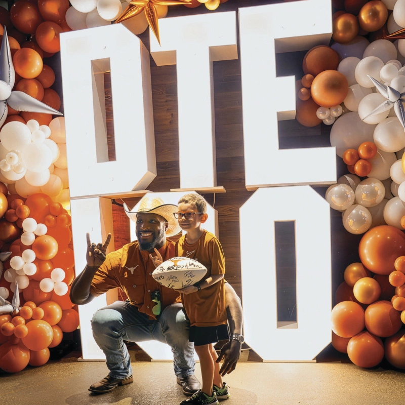 A man in a cowboy hat poses with a child holding a white, signed football. The pair is in front of big, light-box letters that read "DTE 100," surrounded by a burnt-orange and white balloon arch.
