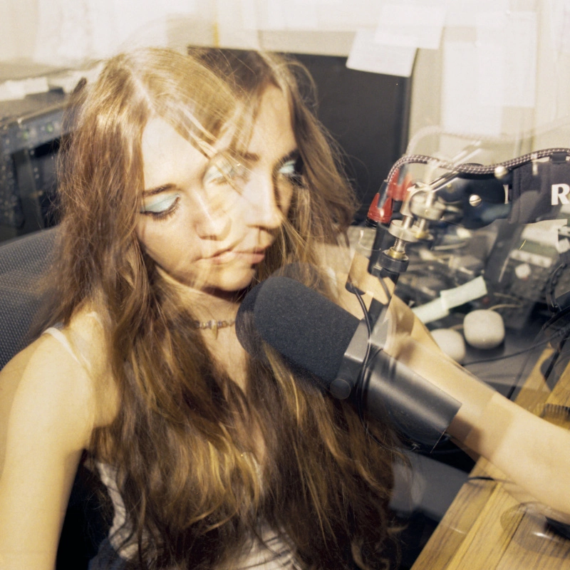 A multiple-exposure picture of a girl with blue eyeshadow and long, fringed hair, sitting in front of a radio microphone.