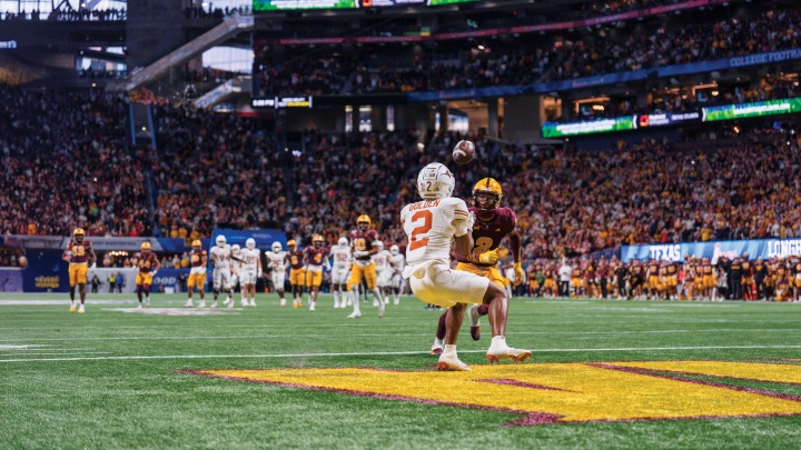 A Longhorn football player in white catches a football ahead of a football player in maroon.