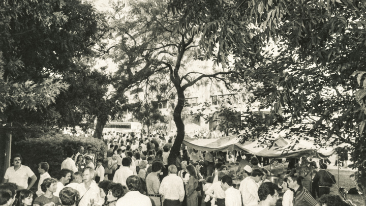 A view of a crowd under the oak trees outside of the Alumni Center, with DKR in the background.