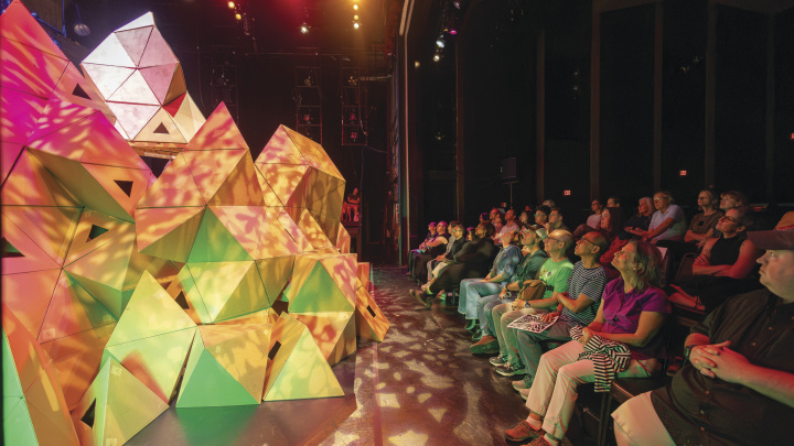A massive geometric set piece protrudes into the watching audience, lit in shades of pink and green.
