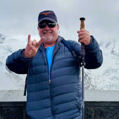 A man in blue jacket does Hook Em Horns hand signal in front of snowy mountains. 
