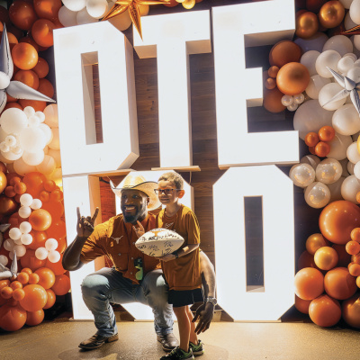 A man in a cowboy hat poses with a child holding a white, signed football. The pair is in front of big, light-box letters that read "DTE 100," surrounded by a burnt-orange and white balloon arch.