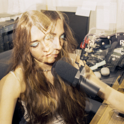 A multiple-exposure picture of a girl with blue eyeshadow and long, fringed hair, sitting in front of a radio microphone.