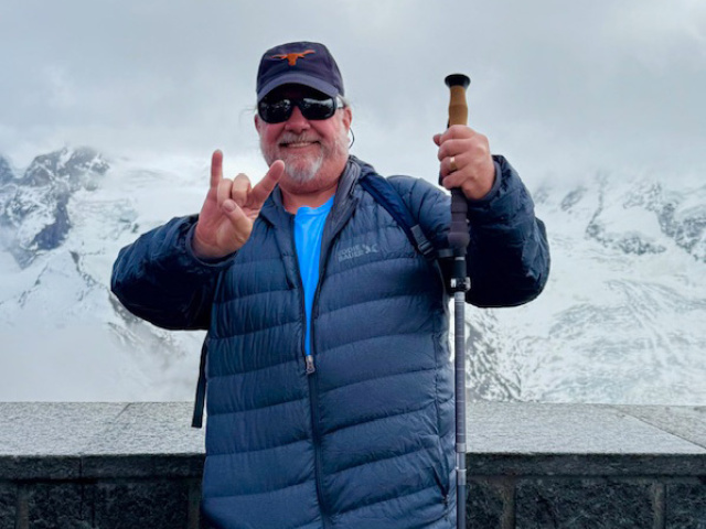 A man in blue jacket does Hook Em Horns hand signal in front of snowy mountains. 