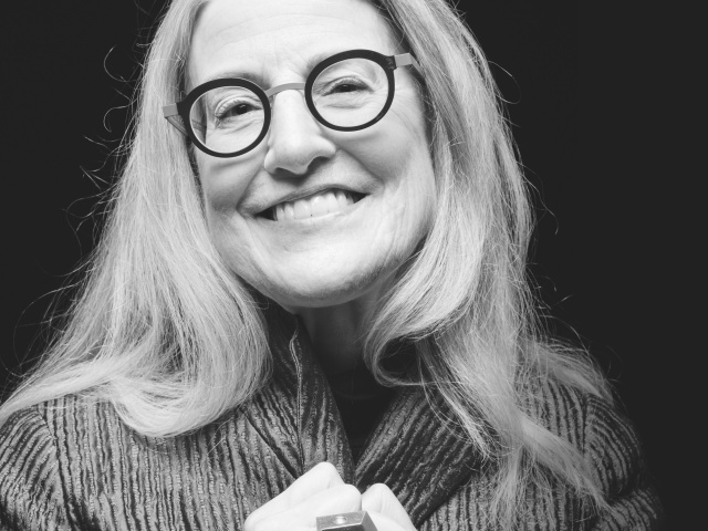 An elderly woman in dark graphic glasses smiles against a black white background.