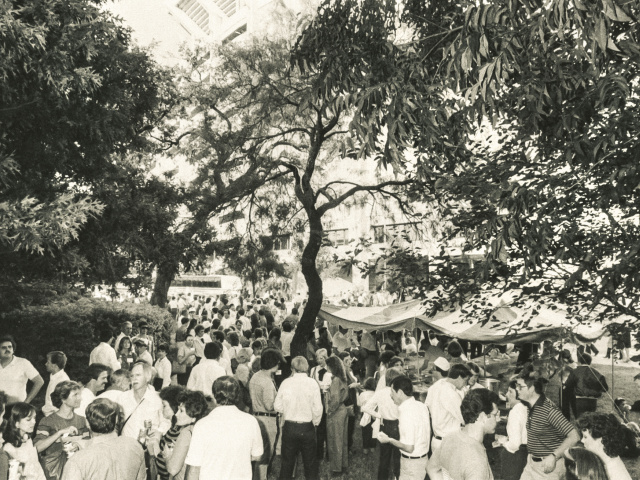 A view of a crowd under the oak trees outside of the Alumni Center, with DKR in the background.