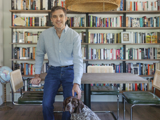 A man stands in jeans and a button down with his dog in front of a shelf full of books.