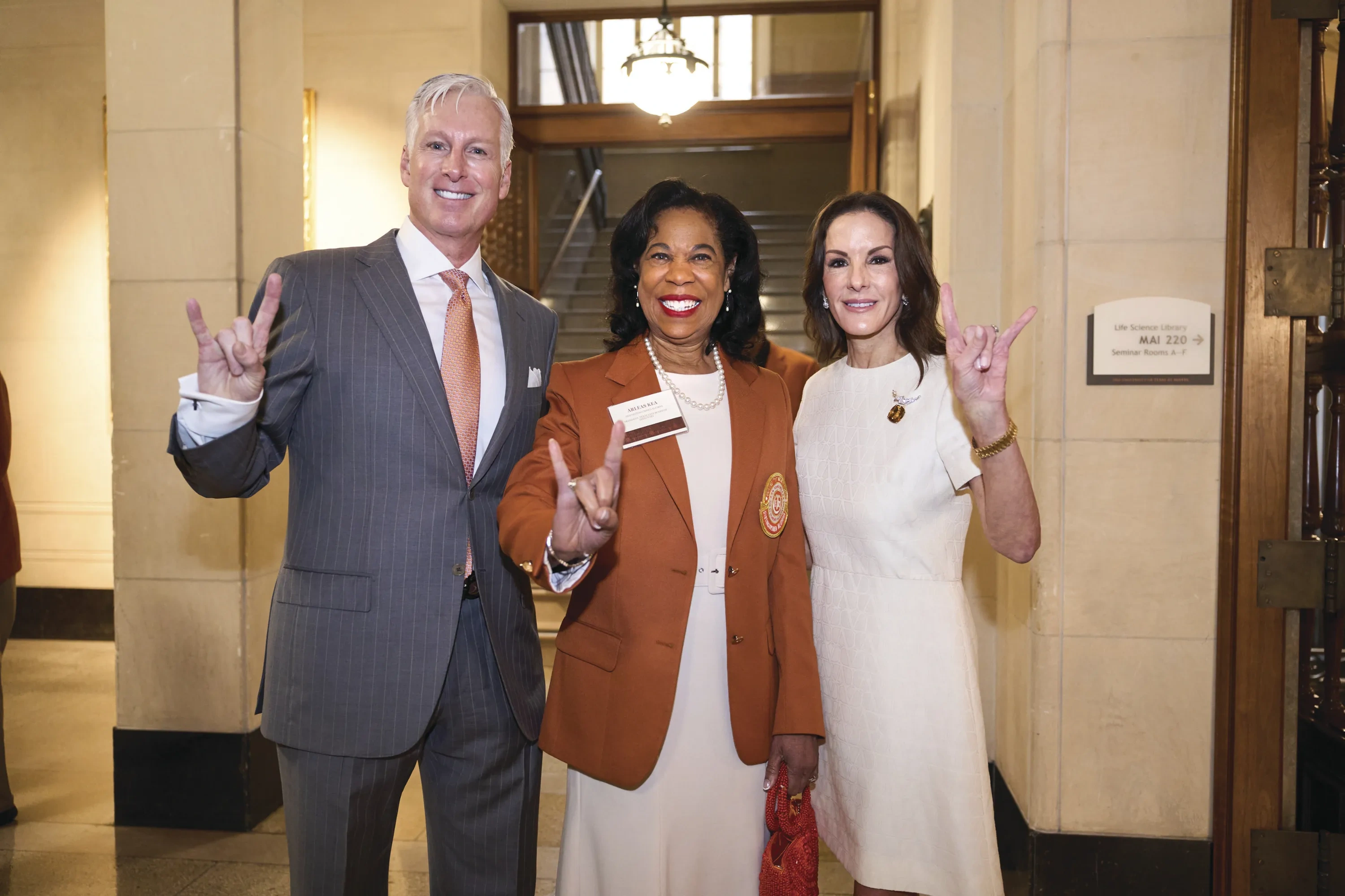 Arleas stands in her burnt-orange Distinguished Alumna blazer, flanked by Robert Jones on the left and a woman in white on the right. All three hook ’em horns.