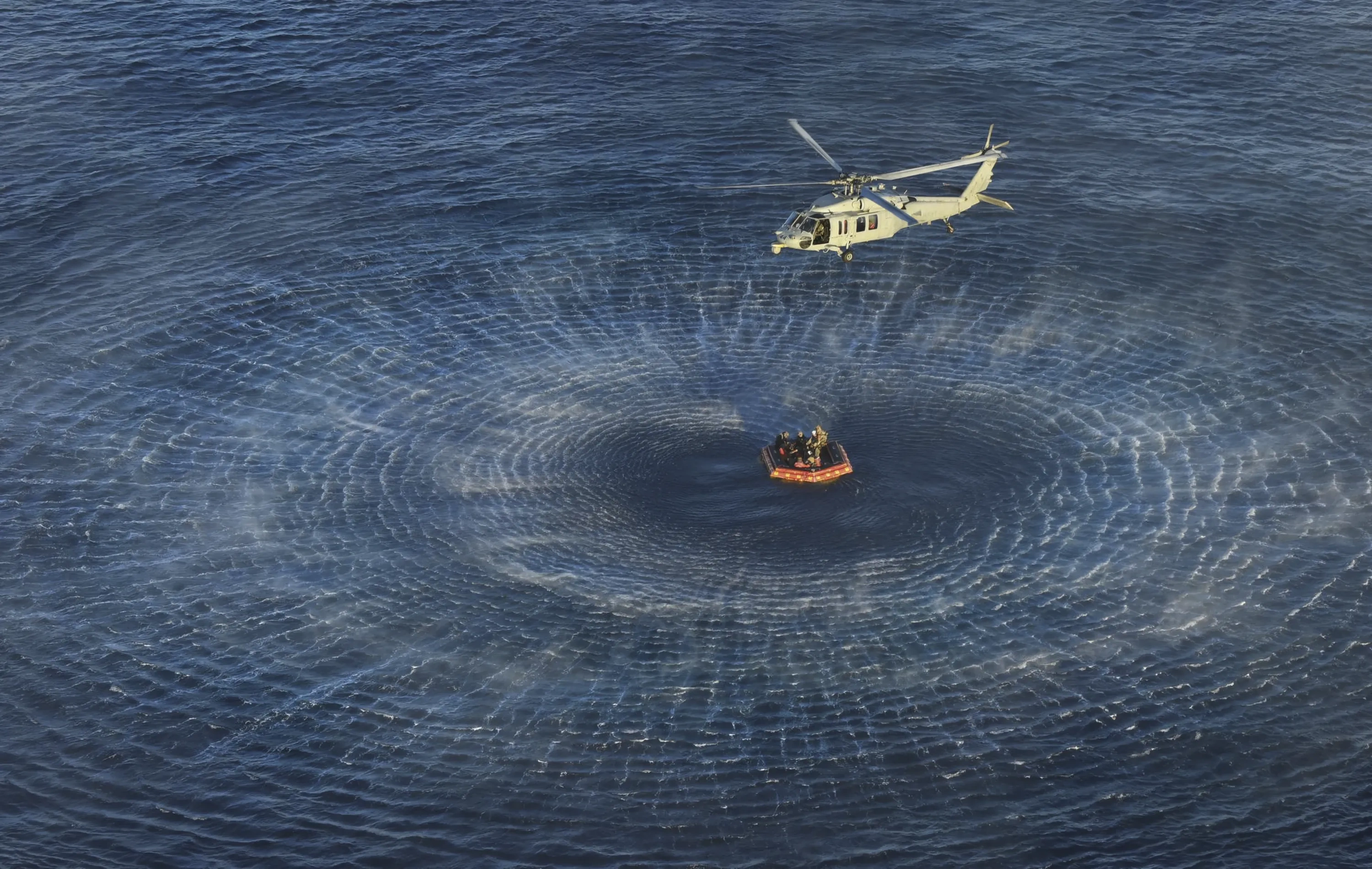 A helicopter hovers over the ocean where a spaceship lander is surrounded by boats.