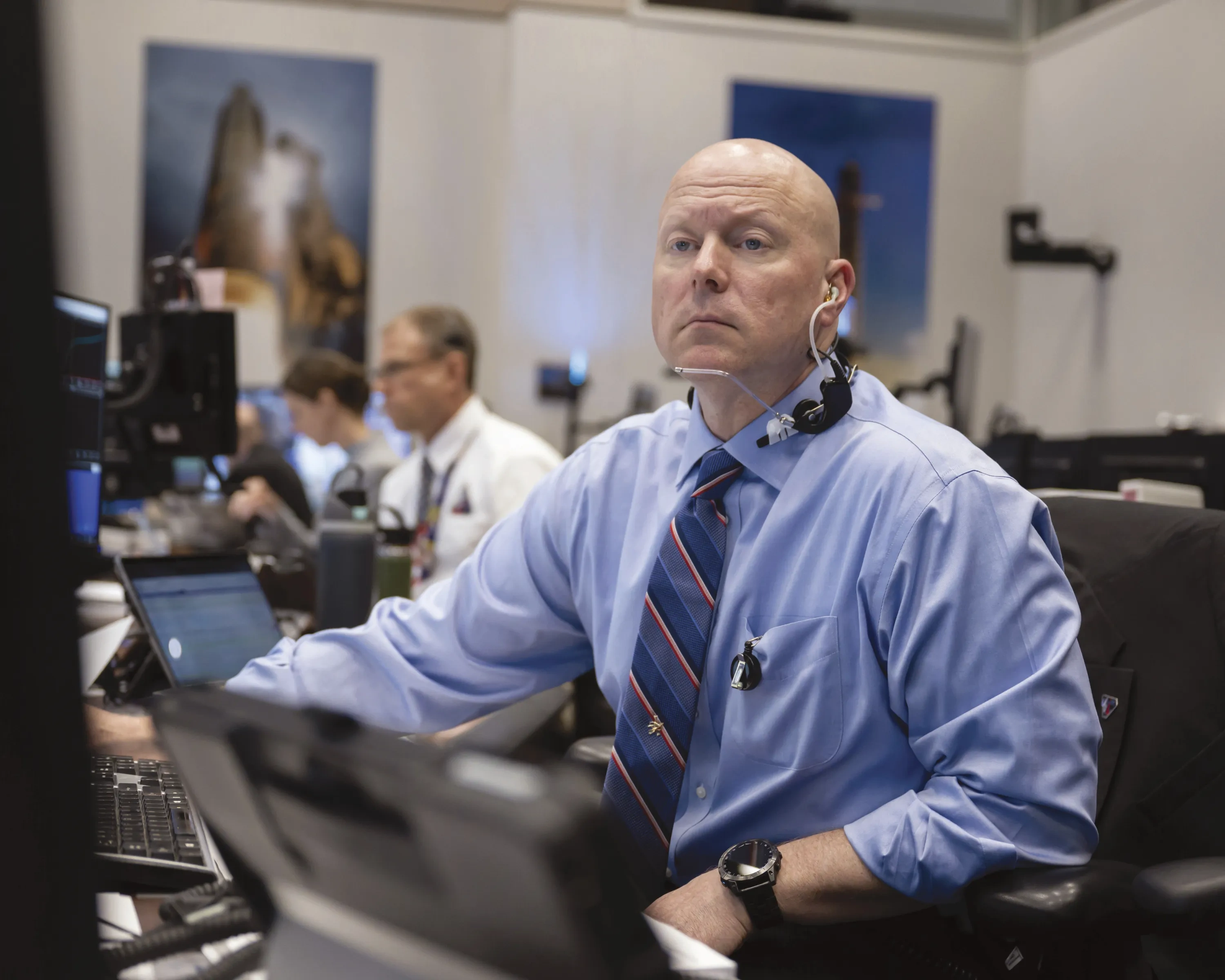 A man in a button down shirt and tie sits at a console in mission control.