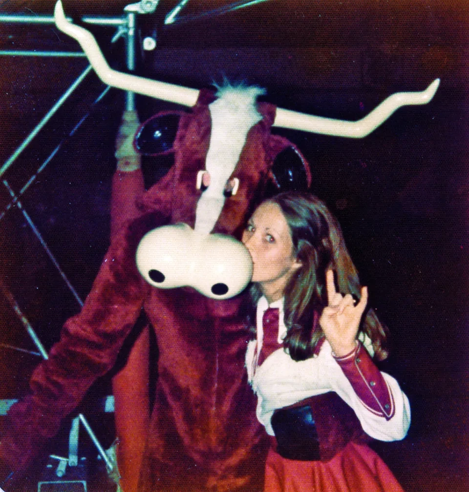 A woman in a cheerleading uniform poses with a retro Longhorn mascot.