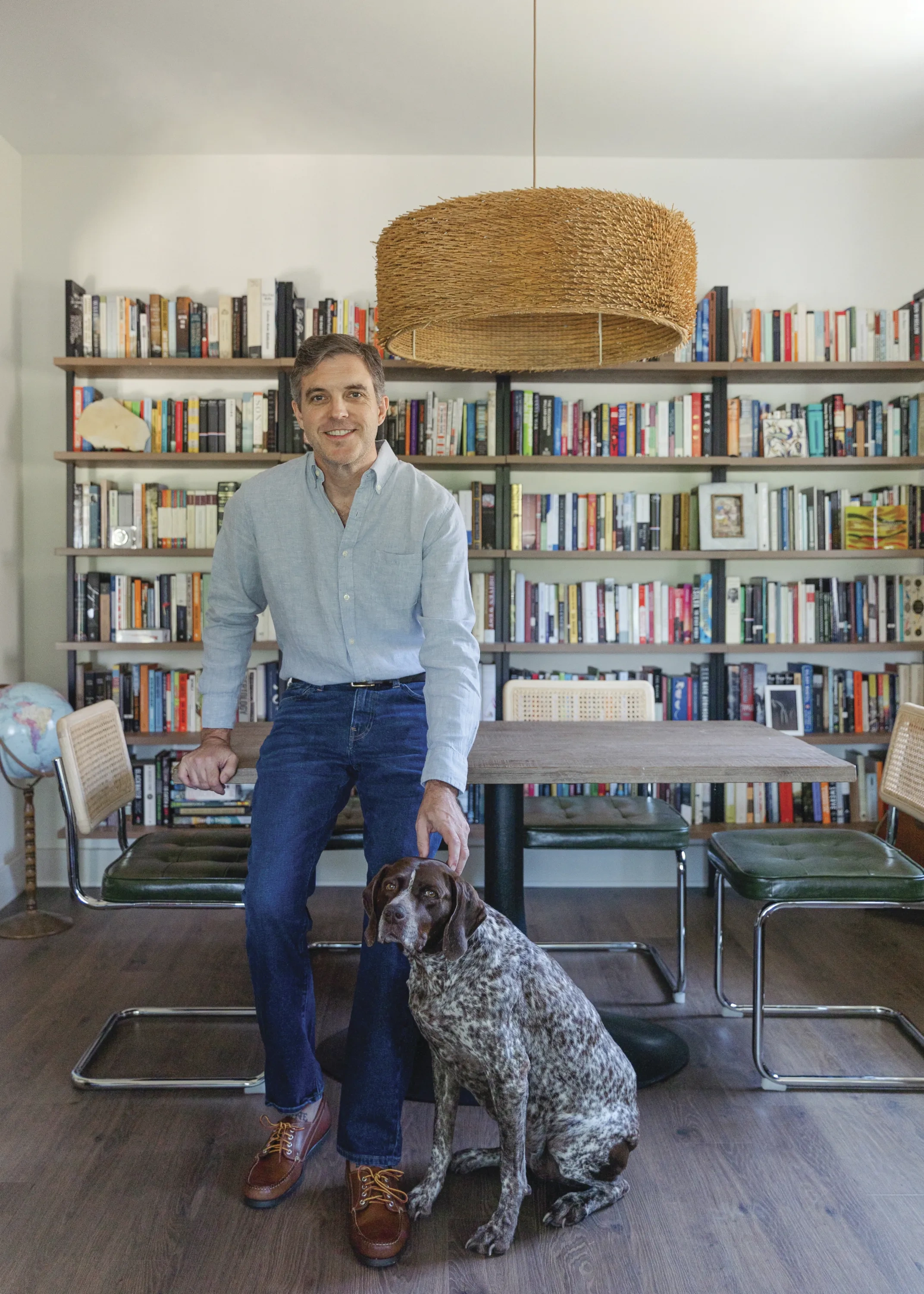 A man stands in jeans and a button down with his dog in front of a shelf full of books.