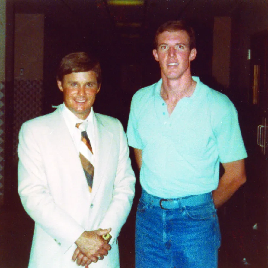 On the left a man in a white suit; on the right a man in a blue short-sleeved polo and jeans.
