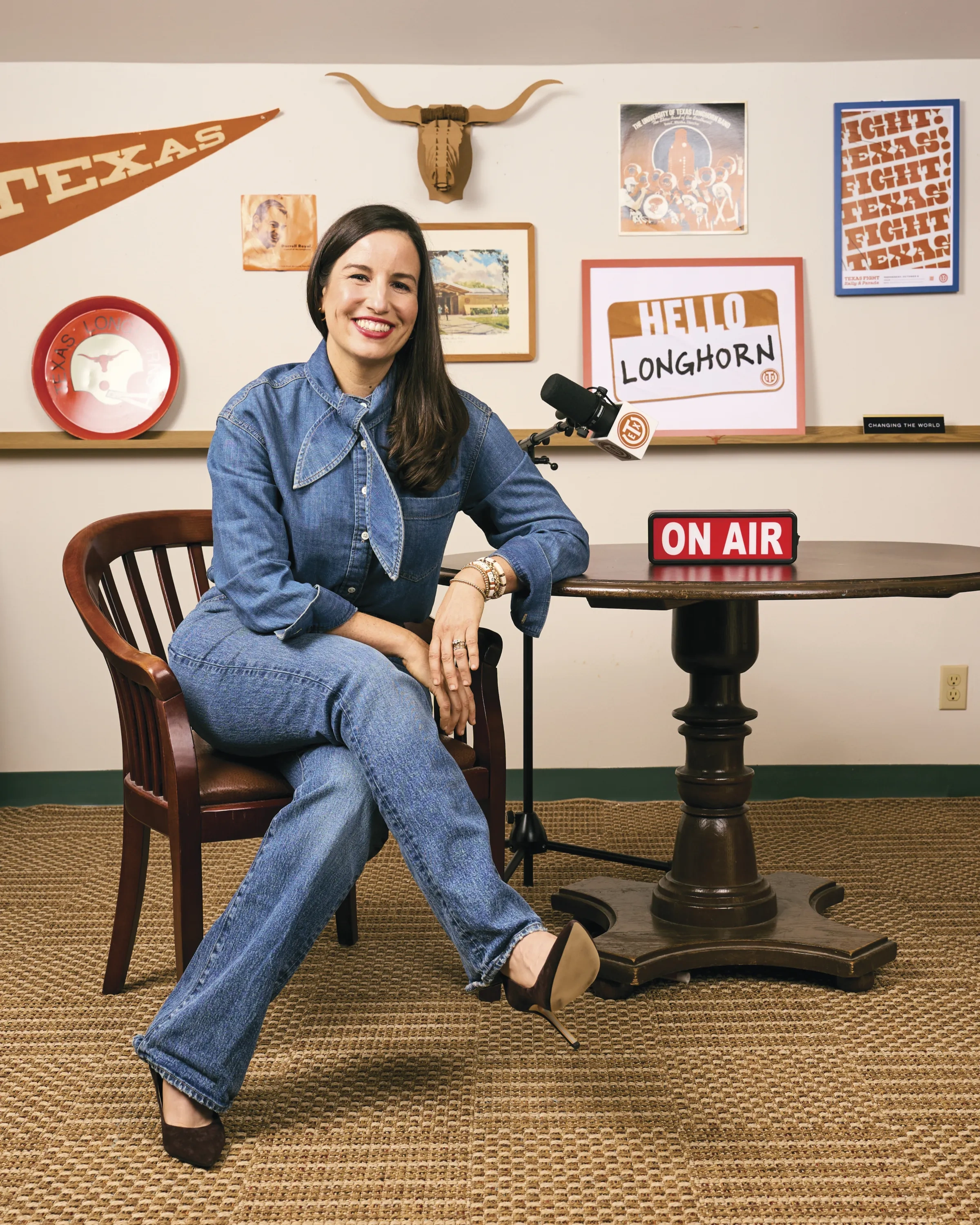 A woman in a denim long-sleeve button down, jeans, and black pumps sits at a table with a red "On Air" sign.
