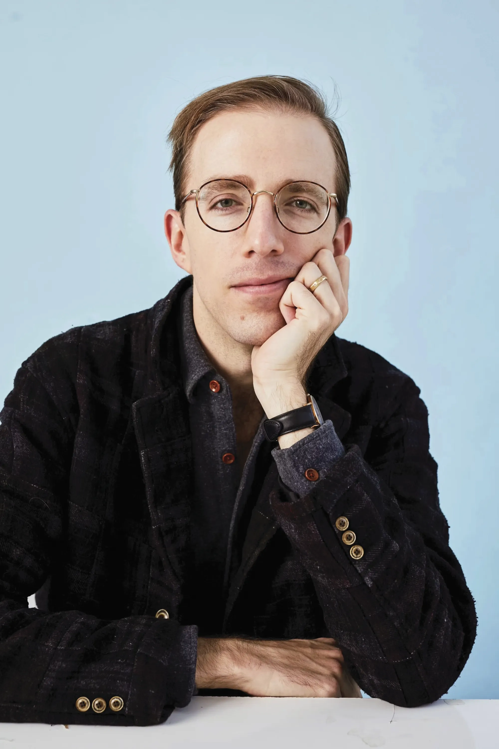 A man in black wire-framed glasses and a black velvet blazer over a grey wool button down props his head on his hands and looks into the camera, against a light blue background.