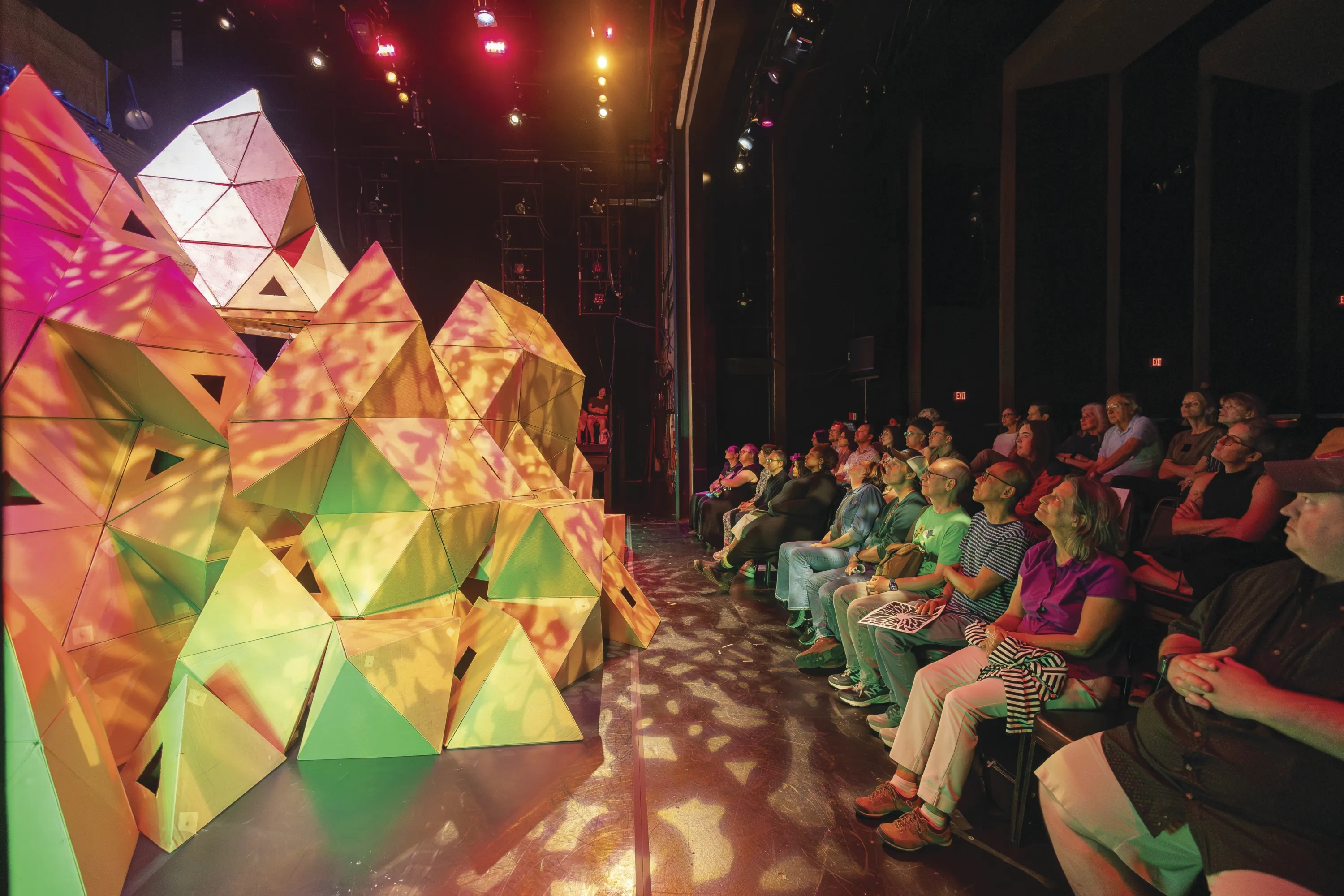 A massive geometric set piece protrudes into the watching audience, lit in shades of pink and green.