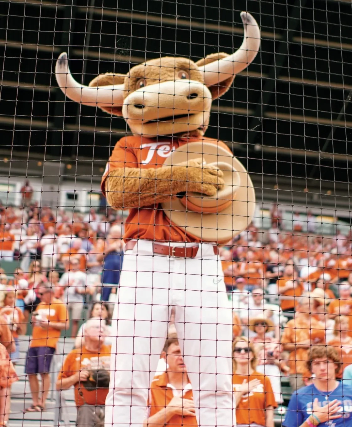 A stuffed Longhorn mascot stands in front of a crowd, holding his cowboy hat over his heart during the National Anthem.