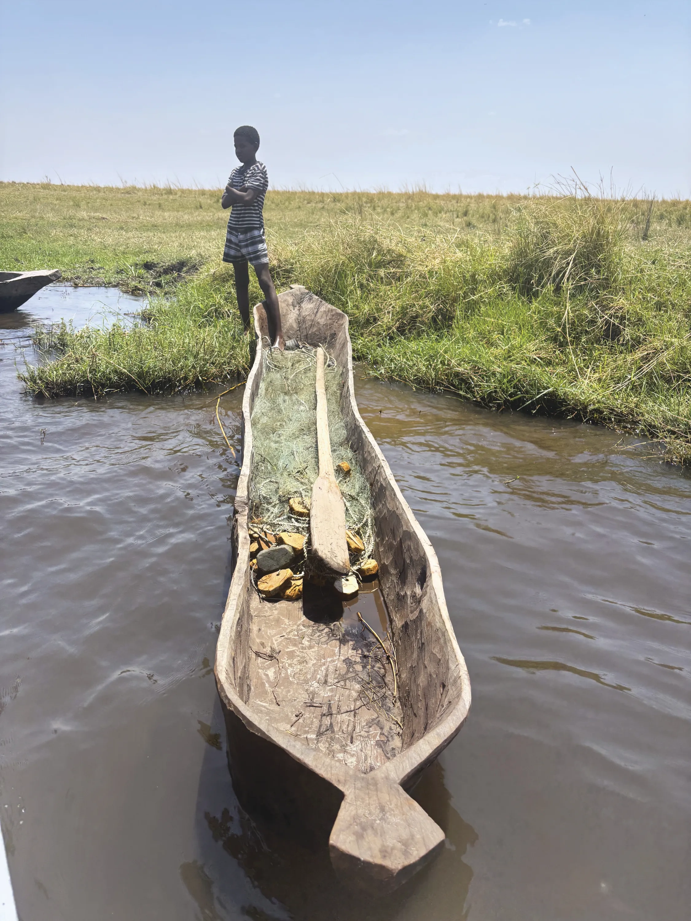 A boy stands on a riverbank in front of a wooden canoe.
