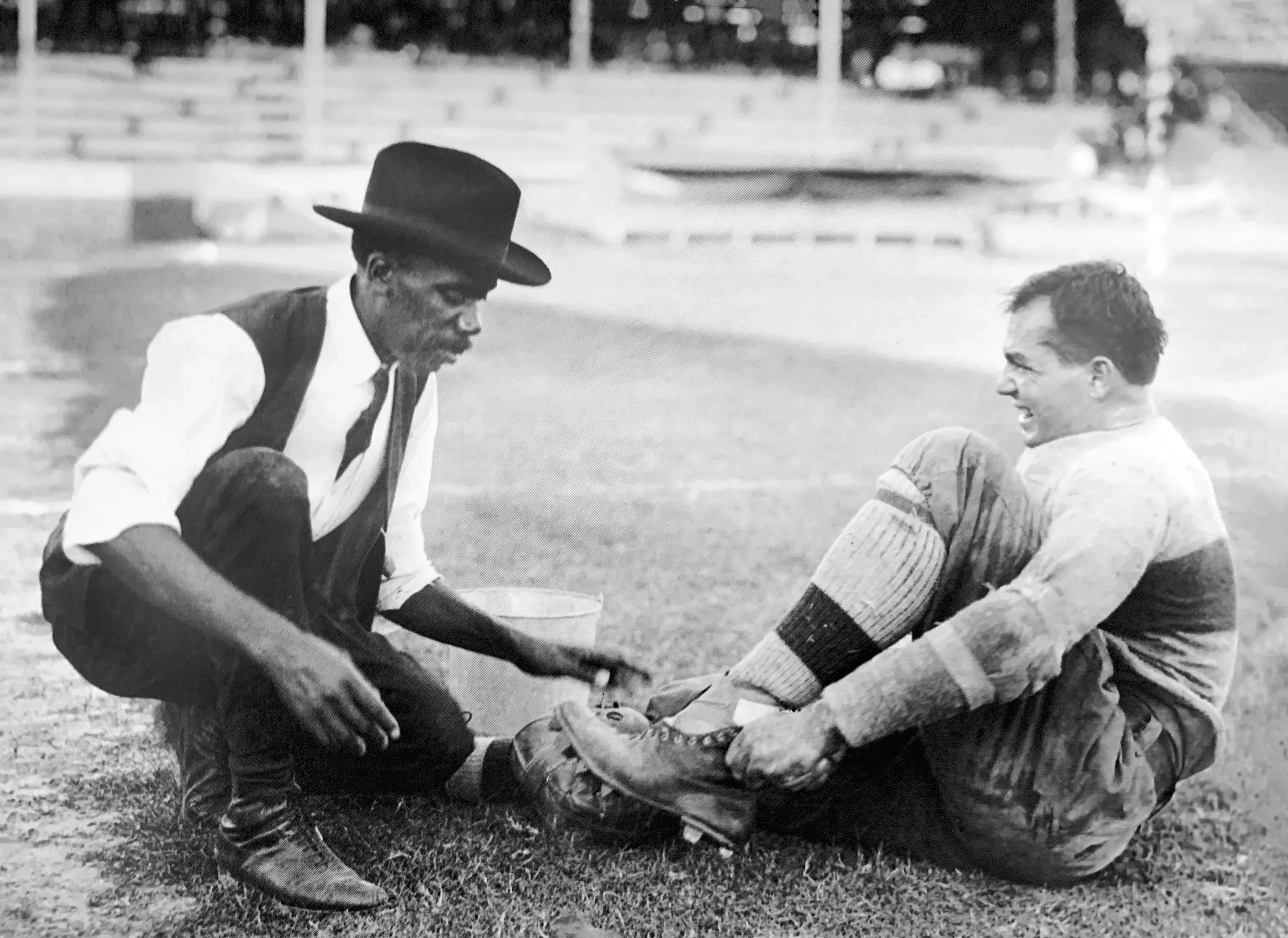 An archival photo in which an older black man in a top hat and vest attends to a young white man who is grimacing in a 1900s-era football uniform.